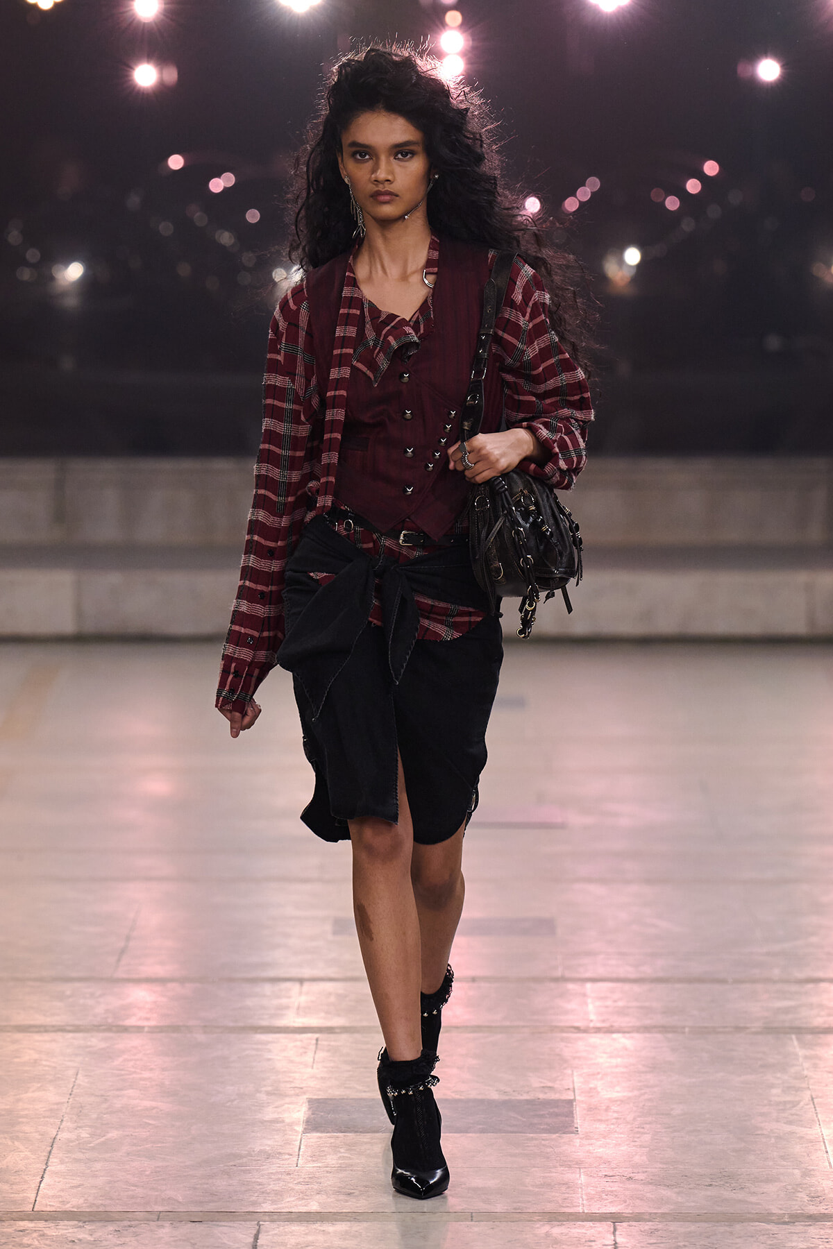 Model walking a nighttime fashion runway in a burgundy plaid shirt over a burgundy vest, with a black wrap skirt and boots, carrying a black handbag.