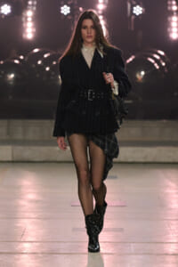 Model on a runway wearing a belted pinstripe blazer with a lace blouse, plaid asymmetrical skirt, fishnet tights and studded black boots.