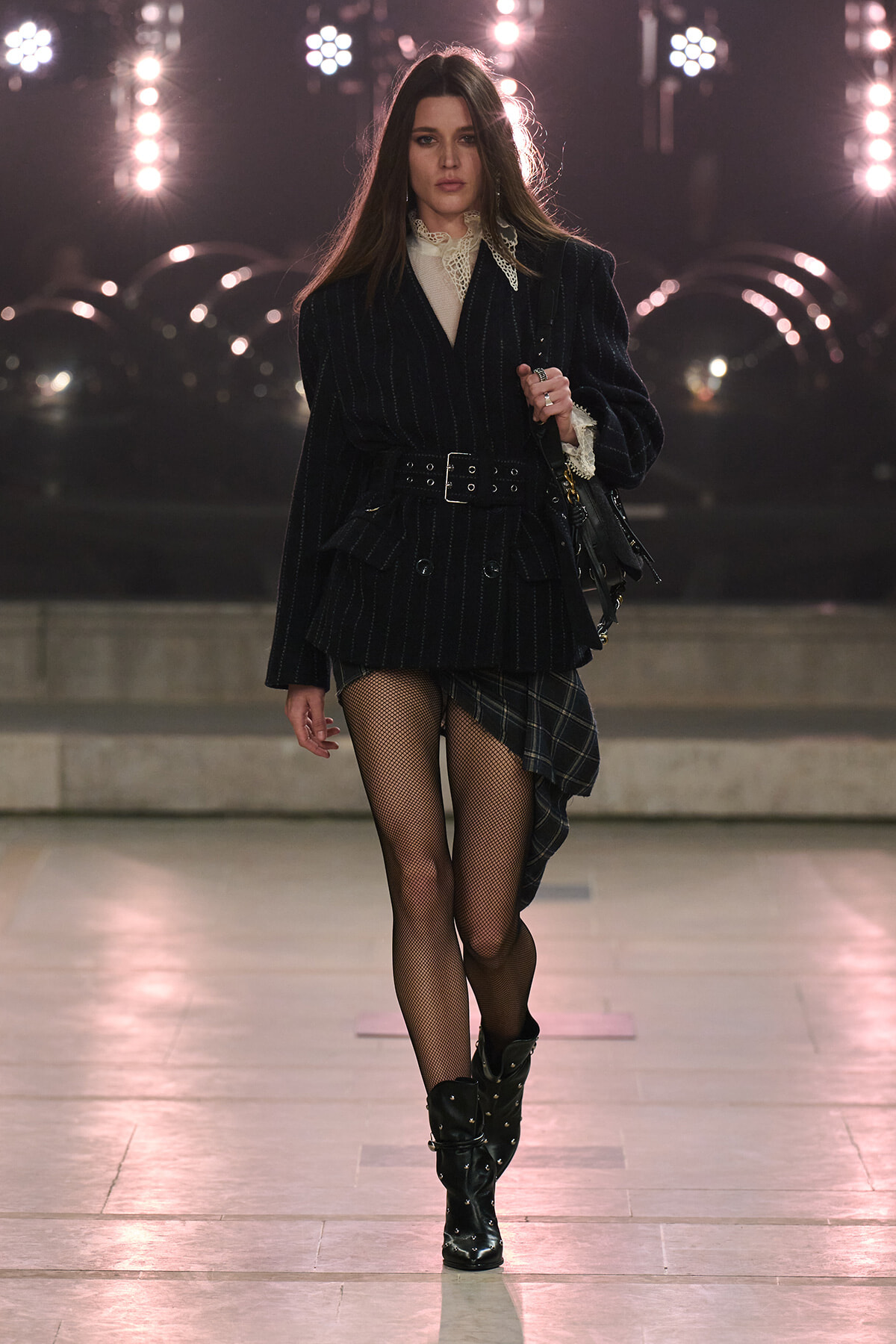 Model on a runway wearing a belted pinstripe blazer with a lace blouse, plaid asymmetrical skirt, fishnet tights and studded black boots.