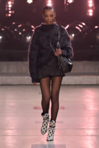 Model on runway wearing a black fuzzy knit sweater, pleated mini skirt, fishnet tights, white polka‑dot ankle boots, and a black crossbody bag.