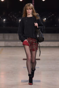Runway model in an oversized black sweater, red plaid mini skirt, fishnet tights, black ankle boots with red toes, and a black shoulder bag.
