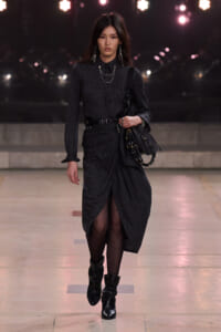 Model walking on a runway in a dark pinstripe dress with a wrapped front, black belt, and chunky boots, carrying a textured black bag with chain accents.