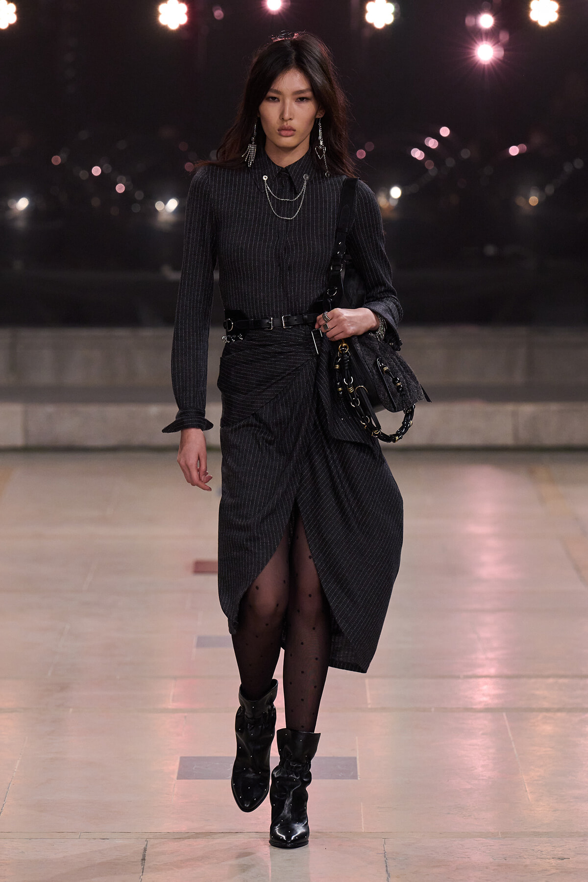 Model walking on a runway in a dark pinstripe dress with a wrapped front, black belt, and chunky boots, carrying a textured black bag with chain accents.