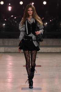 Model on a runway wearing a gray striped shirt with a black layered vest, ruffled mini skirt, lace tights, and black lace-up boots.