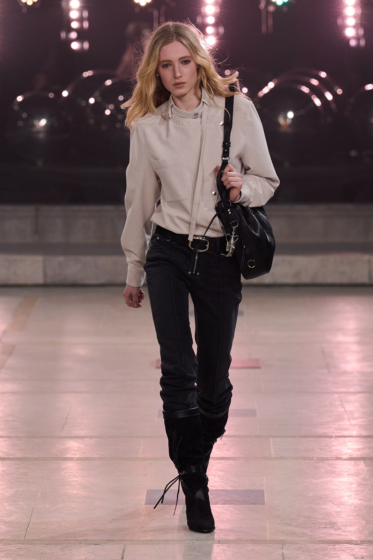 Young woman with blonde hair in a beige jacket, black jeans and knee-high boots walks with a black bag under city lights at night.