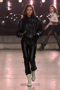Model walks the runway in a black leather oversized jacket and pants with white ankle boots, spotlighted backdrop behind her.
