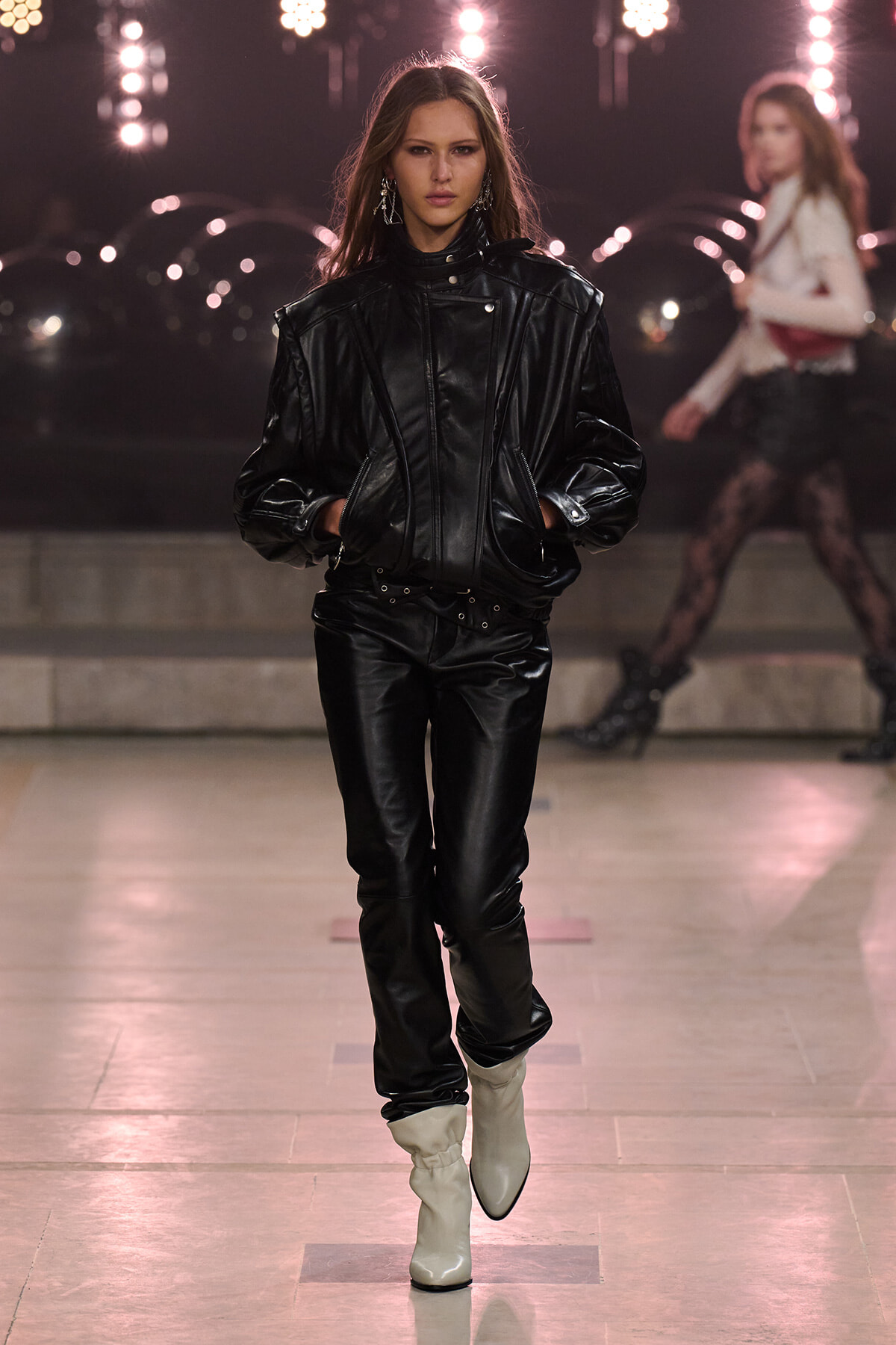 Model walks the runway in a black leather oversized jacket and pants with white ankle boots, spotlighted backdrop behind her.