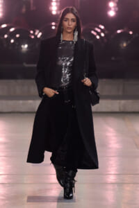 Runway model in a long black coat over a silver sequined top, black pants, and lace-up boots, accessorized with bold dangling earrings.