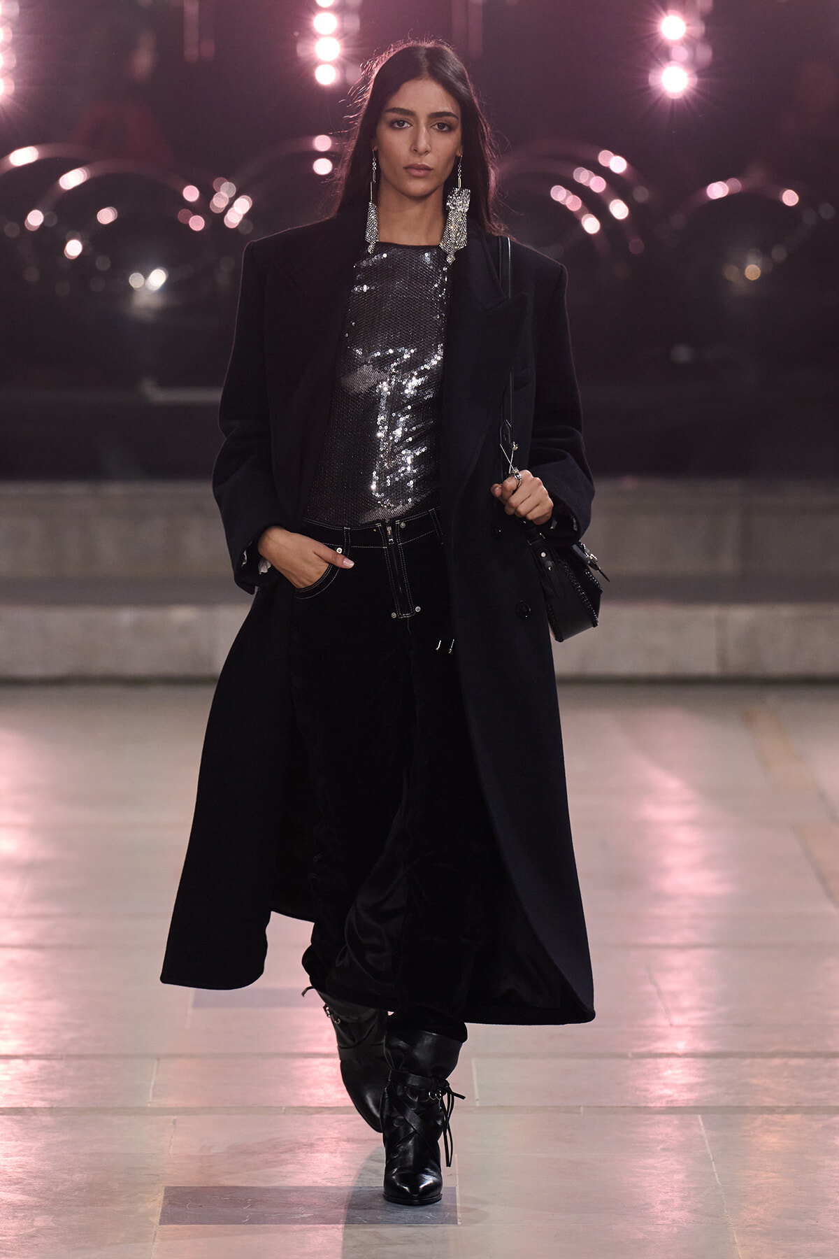 Runway model in a long black coat over a silver sequined top, black pants, and lace-up boots, accessorized with bold dangling earrings.