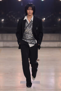 Male model walking a runway in a black suit with a silver sequin top and striped shirt, hands in pockets.