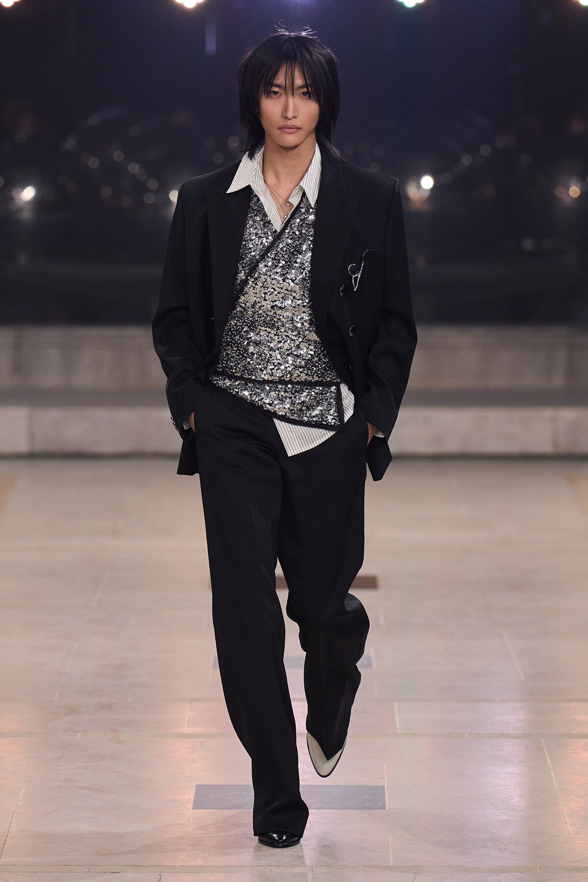 Male model walking a runway in a black suit with a silver sequin top and striped shirt, hands in pockets.