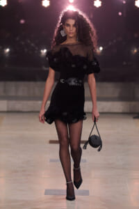 Runway model in a black ruffled dress with a mesh yoke, fishnet stockings, chunky belt, and a round handbag.