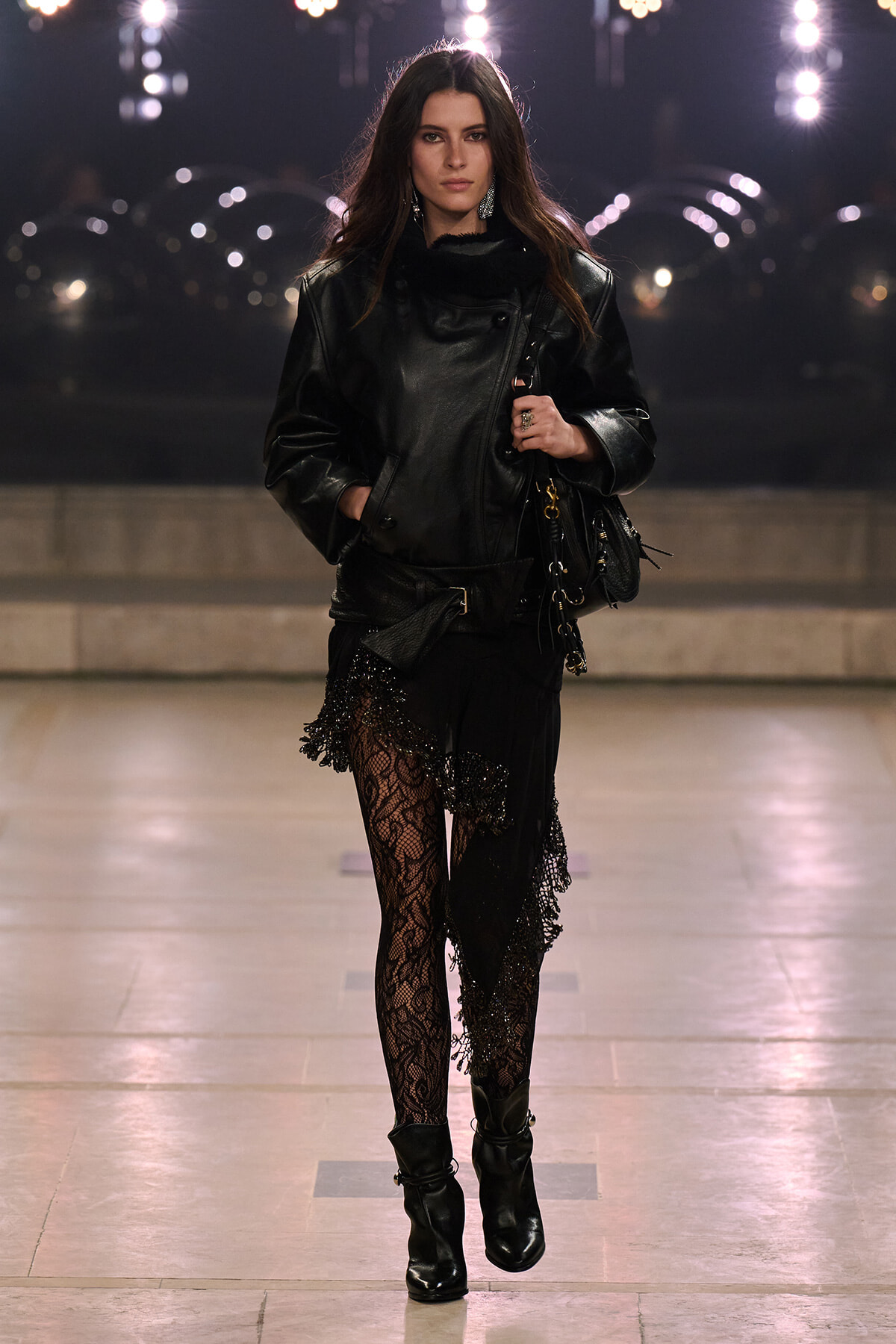 Model walking a runway in a black leather jacket, lace tights, and chunky ankle boots, holding a black bag.