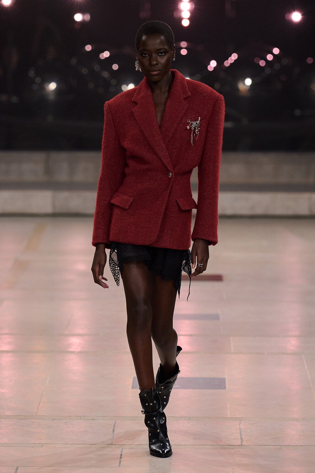 Model on a runway wearing a red textured blazer-styled dress, with black lace shorts and studded black boots.