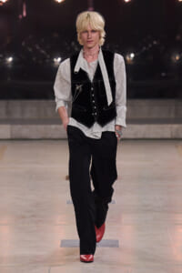 Male model walking a runway in a black velvet vest over a striped shirt, paired with black trousers and red shoes. Accessories include layered necklaces and a chain.
