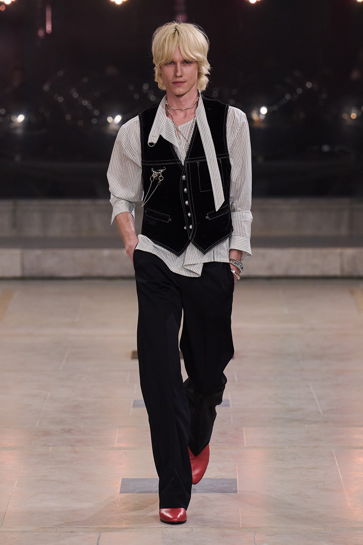 Male model walking a runway in a black velvet vest over a striped shirt, paired with black trousers and red shoes. Accessories include layered necklaces and a chain.