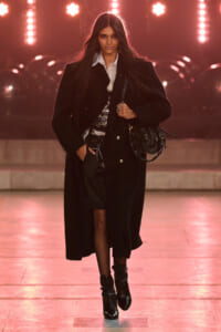 Model walking a runway in a long black coat over a white shirt, sequined top, and black boots, carrying a black handbag as pink stage lights glow behind her.