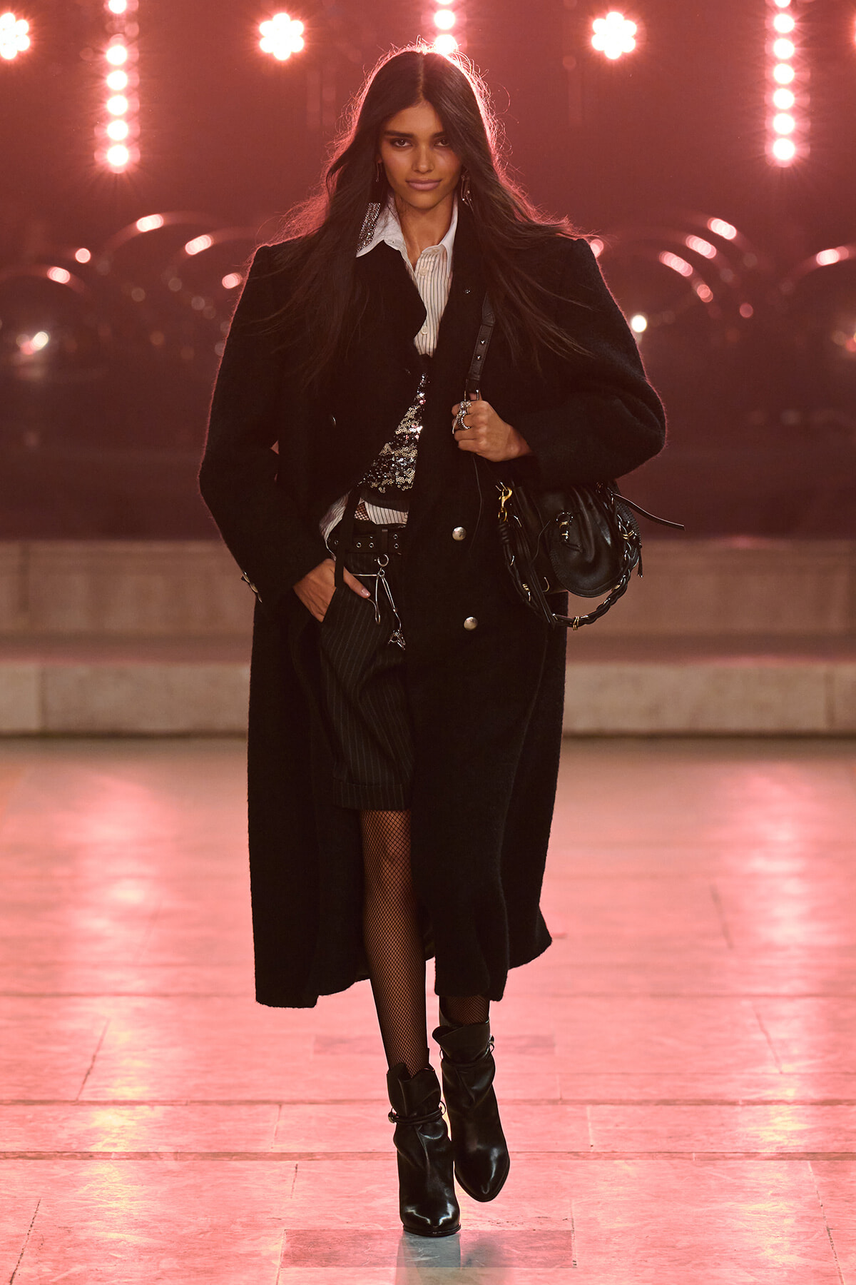Model walking a runway in a long black coat over a white shirt, sequined top, and black boots, carrying a black handbag as pink stage lights glow behind her.