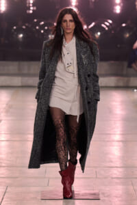 Model walking the runway in a long gray textured coat over a beige mini dress, with black lace tights and red ankle boots.