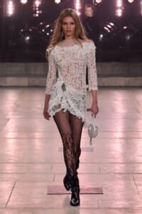 Runway model in a white lace dress with floral pattern, fishnet tights, black studded boots, and a small white circular bag.