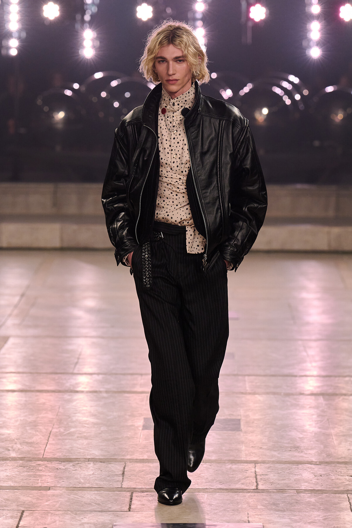 Runway model with blond hair in a black leather jacket, cream polka-dot shirt, and pinstripe pants under bright stage lights.