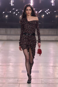 Model walking a runway in an off-shoulder black polka-dot dress with a cinched waist, fishnet tights, black heels, and a small red handbag on a fashion catwalk.
