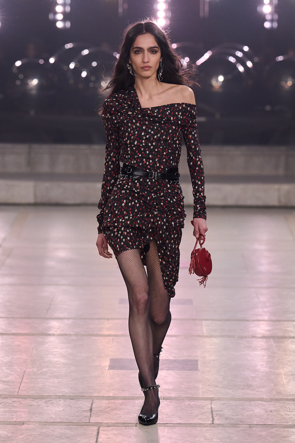 Model walking a runway in an off-shoulder black polka-dot dress with a cinched waist, fishnet tights, black heels, and a small red handbag on a fashion catwalk.