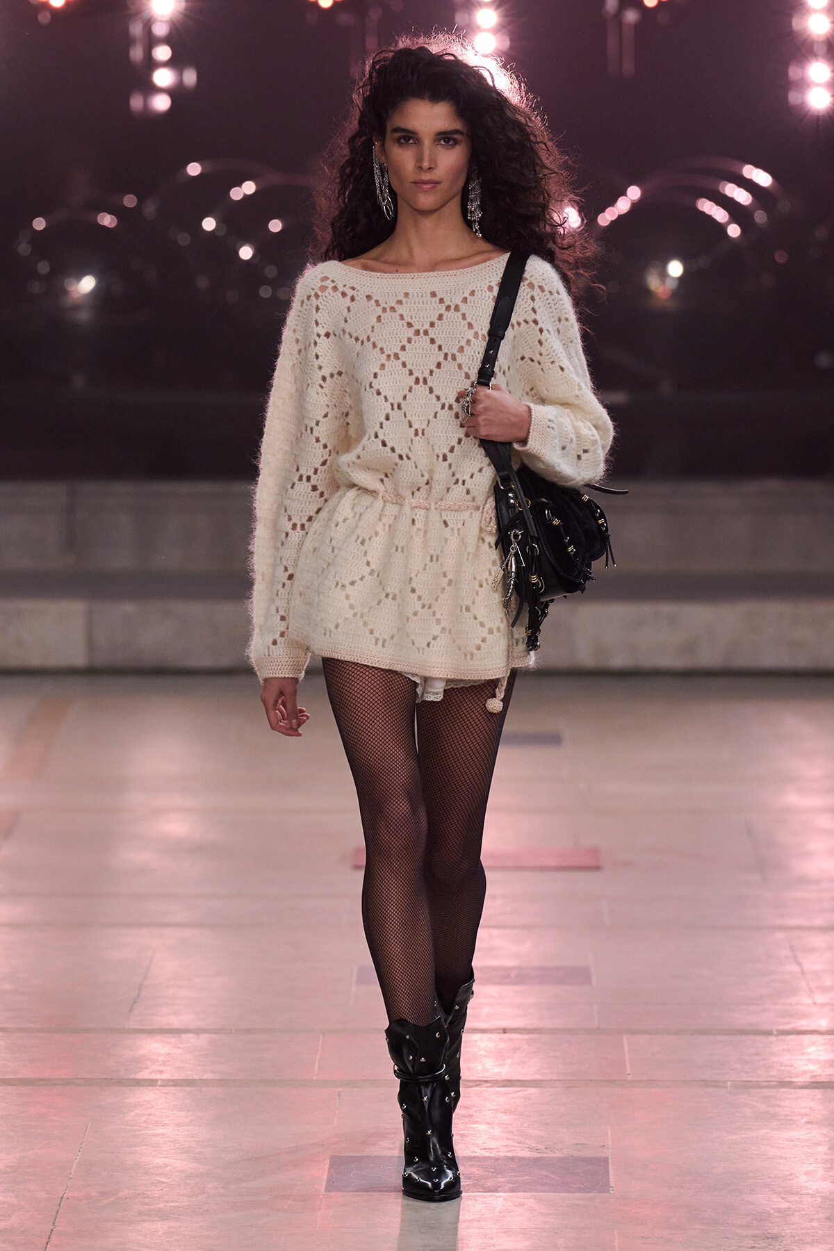 Model walking runway in an oversized cream knitted sweater dress with a belted waist, black fishnet tights, and knee-high black studded boots, carrying a black shoulder bag under pink stage lights.