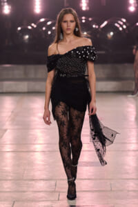 Model on a runway in an off-the-shoulder black top with pearl embellishments, a ruched black skirt, and lace tights.