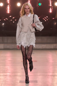 Model walking a runway in a pale striped shirt layered with a beige sleeveless vest, lace-trim shorts, black lace tights, and black pointed heels, carrying a small bag on her shoulder.