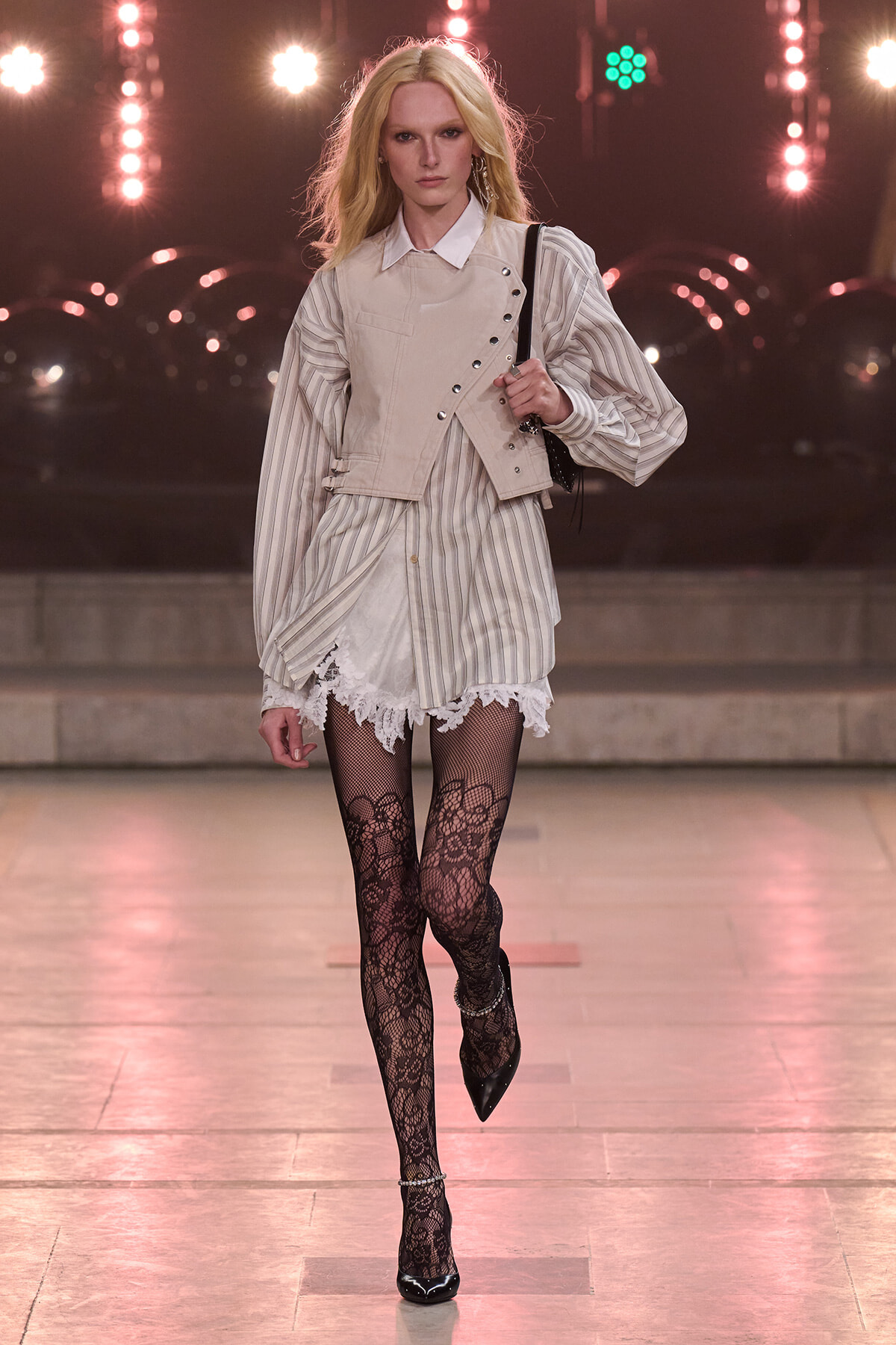 Model walking a runway in a pale striped shirt layered with a beige sleeveless vest, lace-trim shorts, black lace tights, and black pointed heels, carrying a small bag on her shoulder.