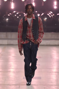 Male model walking a fashion runway at night, wearing a red plaid shirt, dark vest, grey tee, loose jeans and black boots.