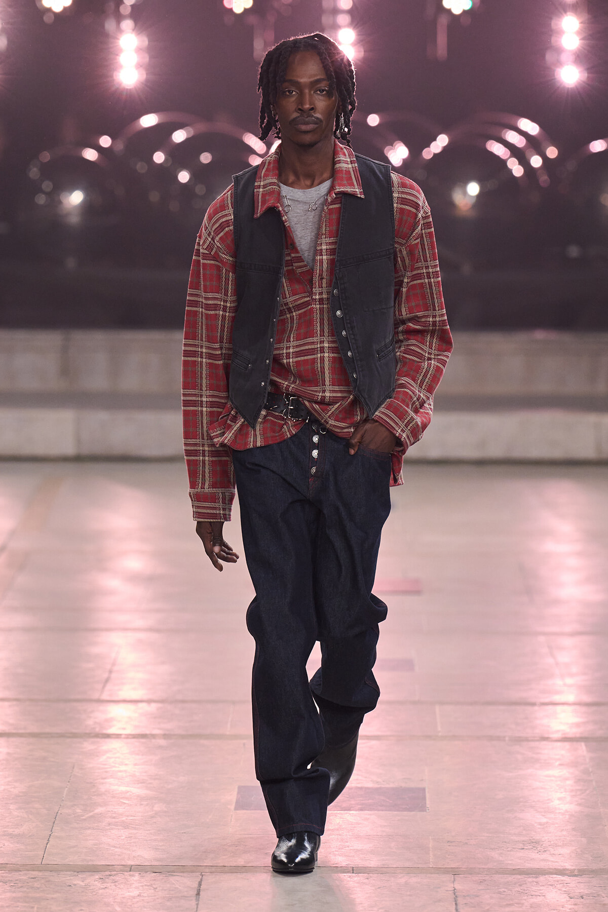 Male model walking a fashion runway at night, wearing a red plaid shirt, dark vest, grey tee, loose jeans and black boots.
