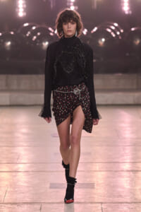 Female model walking a runway in a black, sequined blouse with layered necklaces and a black-dotted wrap skirt with a silver belt buckle; black socks and red-toe heeled shoes visible.