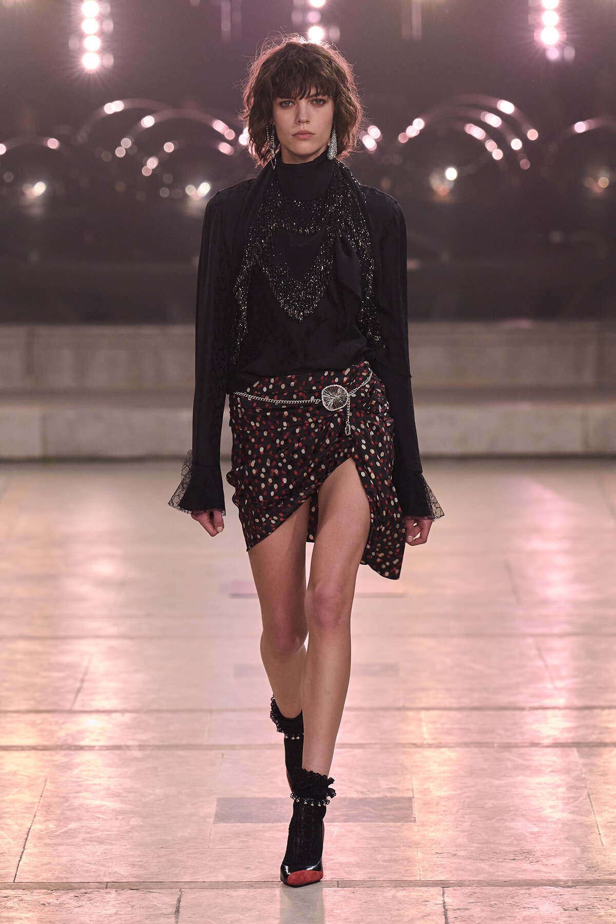 Female model walking a runway in a black, sequined blouse with layered necklaces and a black-dotted wrap skirt with a silver belt buckle; black socks and red-toe heeled shoes visible.