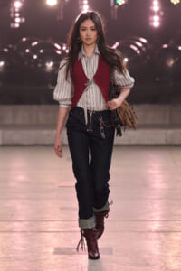 Female model walking a fashion runway in a striped button-down, burgundy vest, dark jeans with cuffs, and burgundy fringe boots, carrying a brown bag.