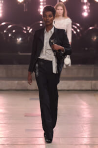 Runway model in a dark pinstripe suit with a white striped blouse, carrying a black handbag on the catwalk.