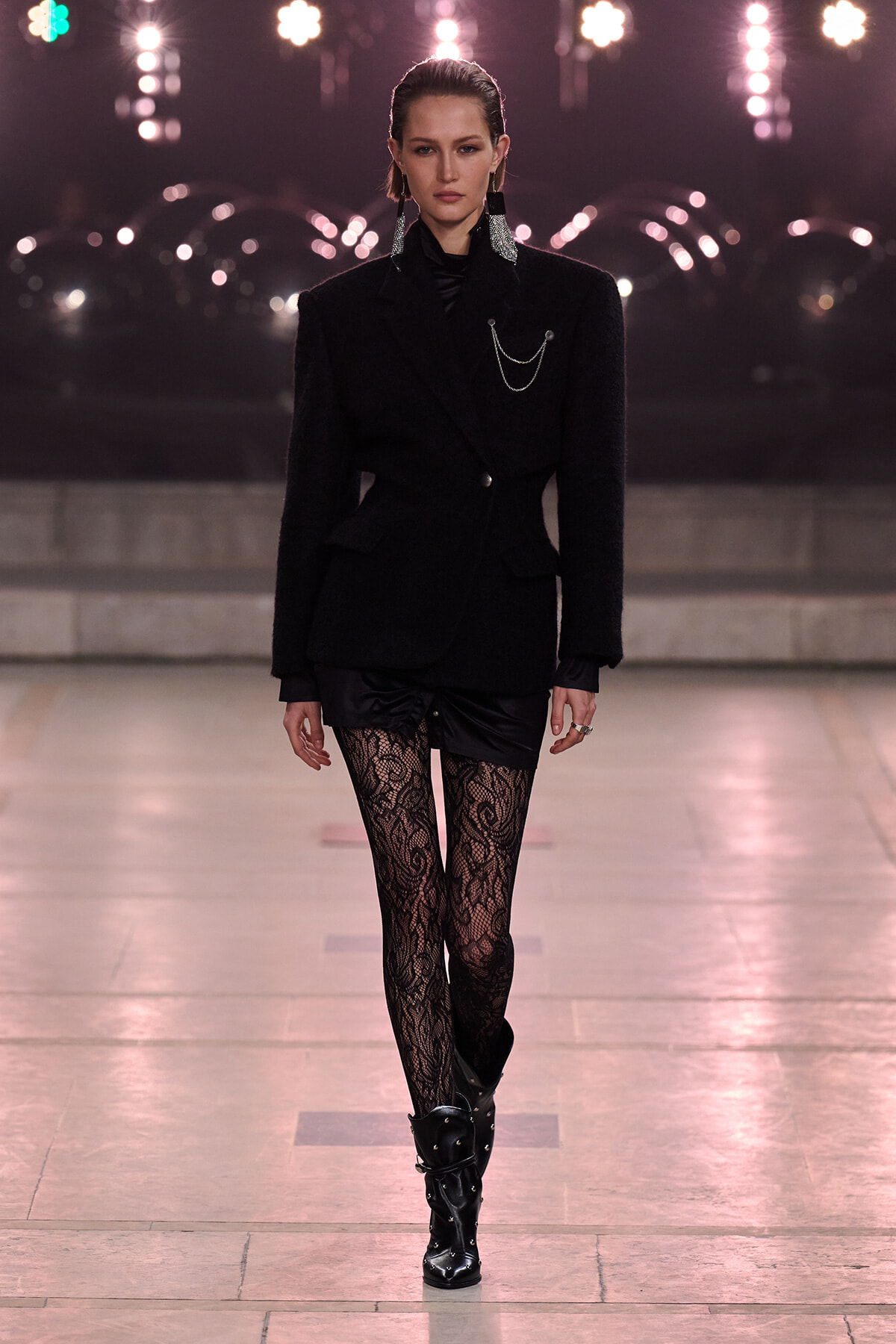 Runway model in an all-black look: tailored blazer with chain detail, lacy tights, and studded knee-high boots on a fashion catwalk.