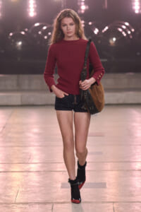 Runway model walking in a red knit sweater, dark shorts, black ankle boots, and a brown shoulder bag on a fashion show catwalk.