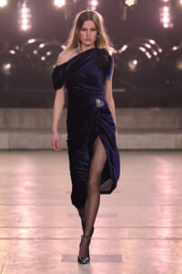 Model on a runway wearing a dark blue velvet one-shoulder gown with draped bodice and a high slit, paired with fishnet tights and black heels.