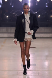 Model walking the runway in a black blazer over a sheer lace blouse and mini lace skirt, black ankle boots with embellishments