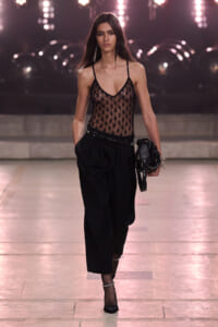 Model walking a runway in a black lace mesh bodysuit, black high-waisted pleated pants, fishnet stockings, black heels, and a black clutch with chains.