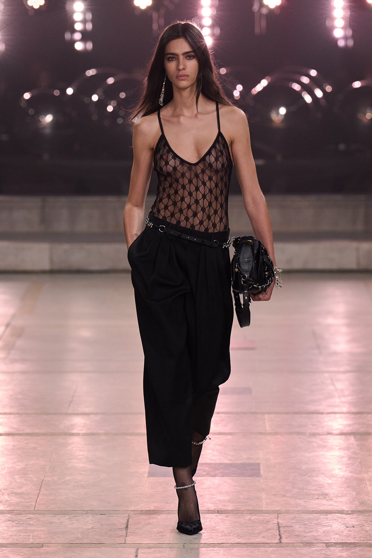 Model walking a runway in a black lace mesh bodysuit, black high-waisted pleated pants, fishnet stockings, black heels, and a black clutch with chains.
