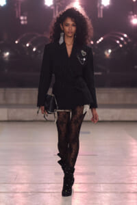 Female model walks a runway in a black pinstripe double-breasted blazer dress, lace tights, studded boots, and statement earrings against a bright backstage backdrop.