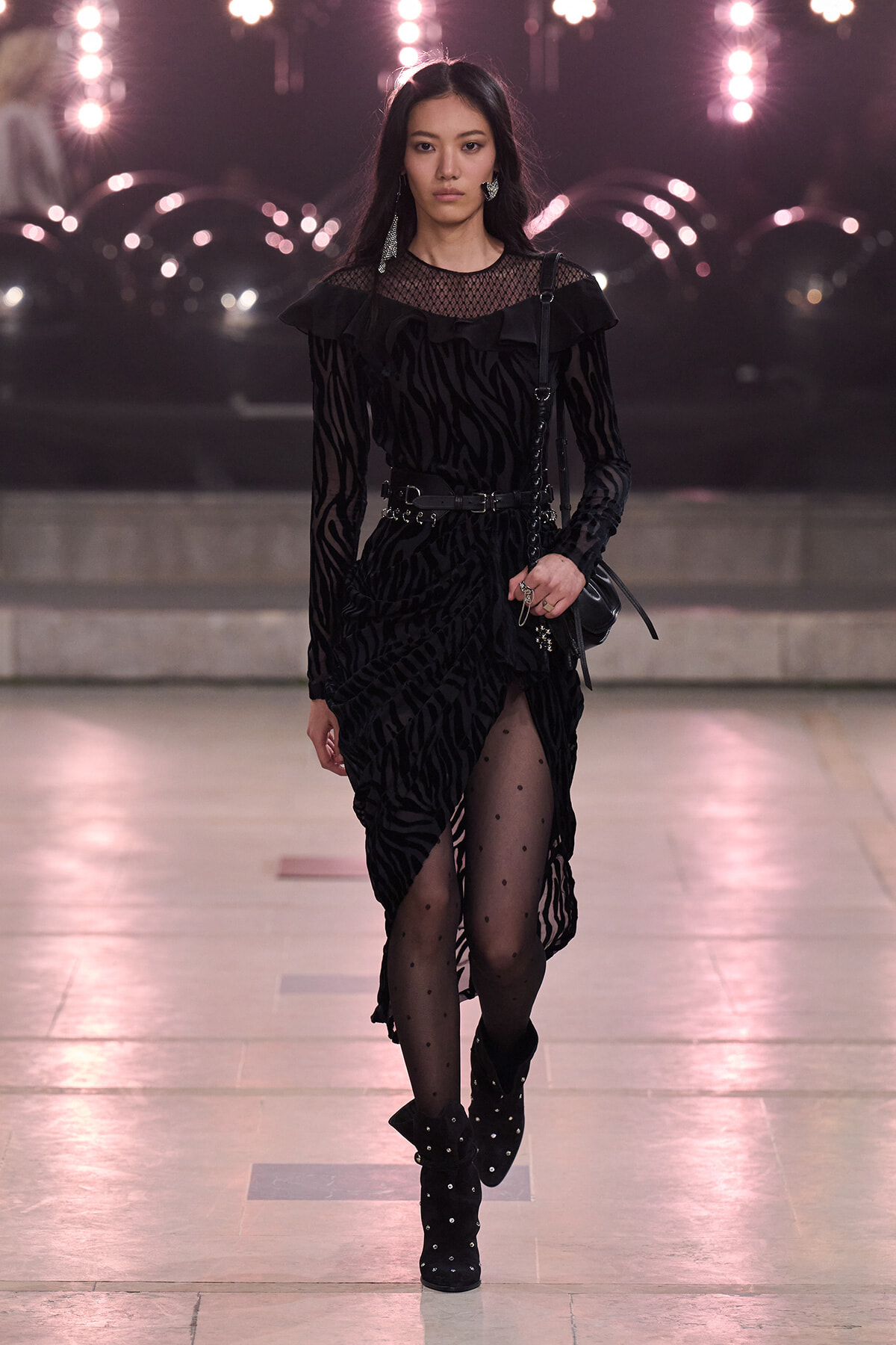 Model walks a fashion runway in a black zebra-patterned dress with sheer yoke, ruffles, a belt, and dotted tights paired with studded boots.