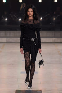 Model walking the runway in a black polka-dot, off-shoulder dress with sheer neckline and lace tights, carrying a small black bag with a silver belt.”,
