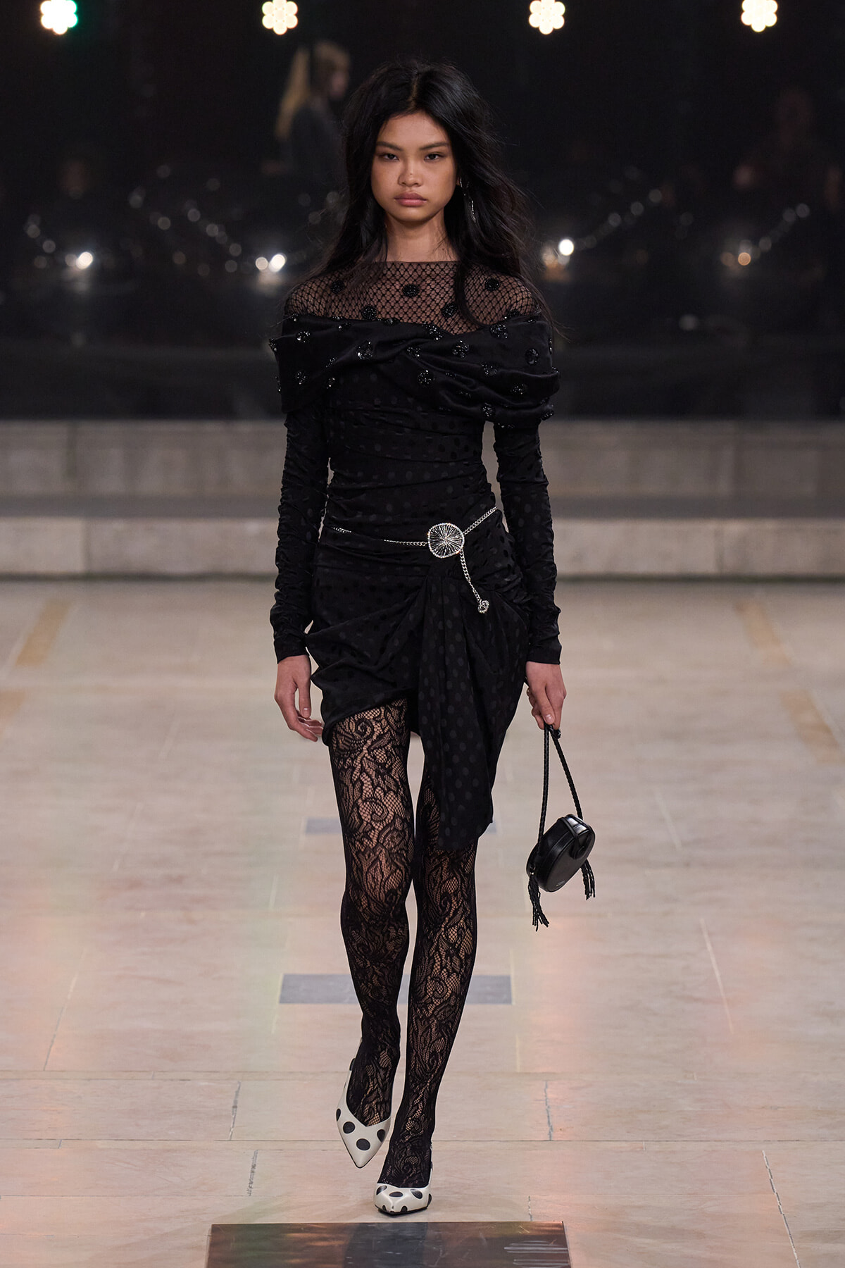 Model walking the runway in a black polka-dot, off-shoulder dress with sheer neckline and lace tights, carrying a small black bag with a silver belt.”,