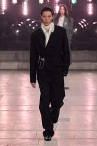 Male model on a fashion runway in a black pinstripe suit with a fringed white scarf and silver chains, under bright stage lights.