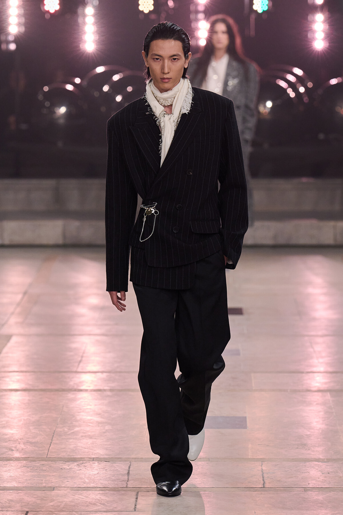 Male model on a fashion runway in a black pinstripe suit with a fringed white scarf and silver chains, under bright stage lights.