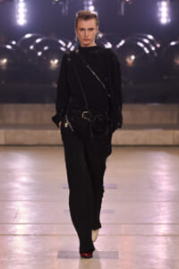 Model walking a runway in a black outfit with a crossbody chain and small black purse, spotlighted on stage.